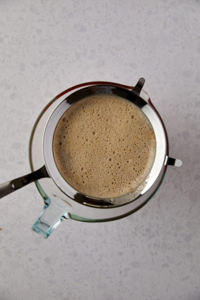 A glass measuring cup filled with frothy brown liquid on a speckled gray surface. A metal sieve with a handle rests on the cup, suggesting the liquid is being strained.