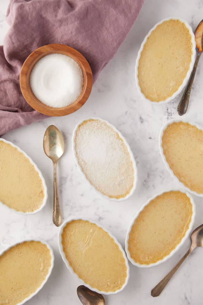 A variety of oval ramekins filled with crème brûlée are arranged on a marble surface. A wooden bowl of sugar and several spoons are placed nearby, and a mauve fabric napkin is partially visible in the corner.