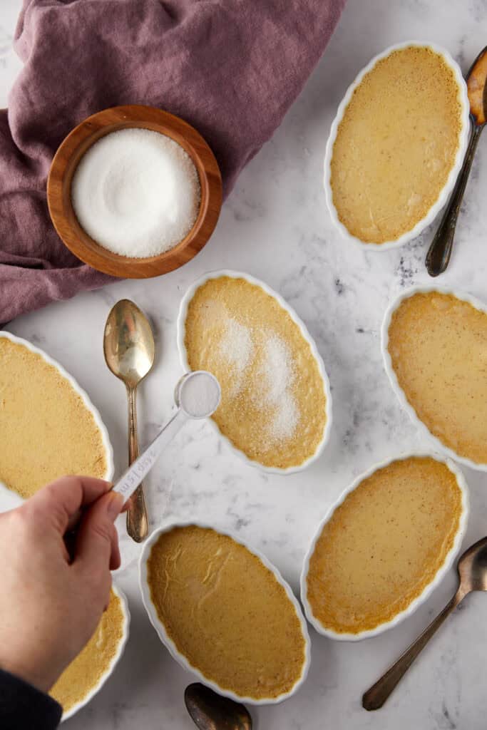 Various ramekins filled with creme brulee are arranged on a marble countertop. A hand is sprinkling sugar onto one dessert, with a bowl of sugar and a purple cloth nearby. Several spoons are placed around the ramekins.