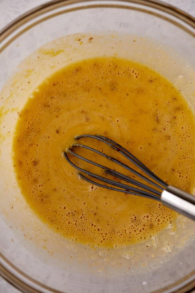 A glass bowl containing a whisk and a yellowish-brown mixture, likely a blend of egg and spices, is shown. The mixture appears smooth with tiny air bubbles on the surface, and the whisk rests inside the bowl.