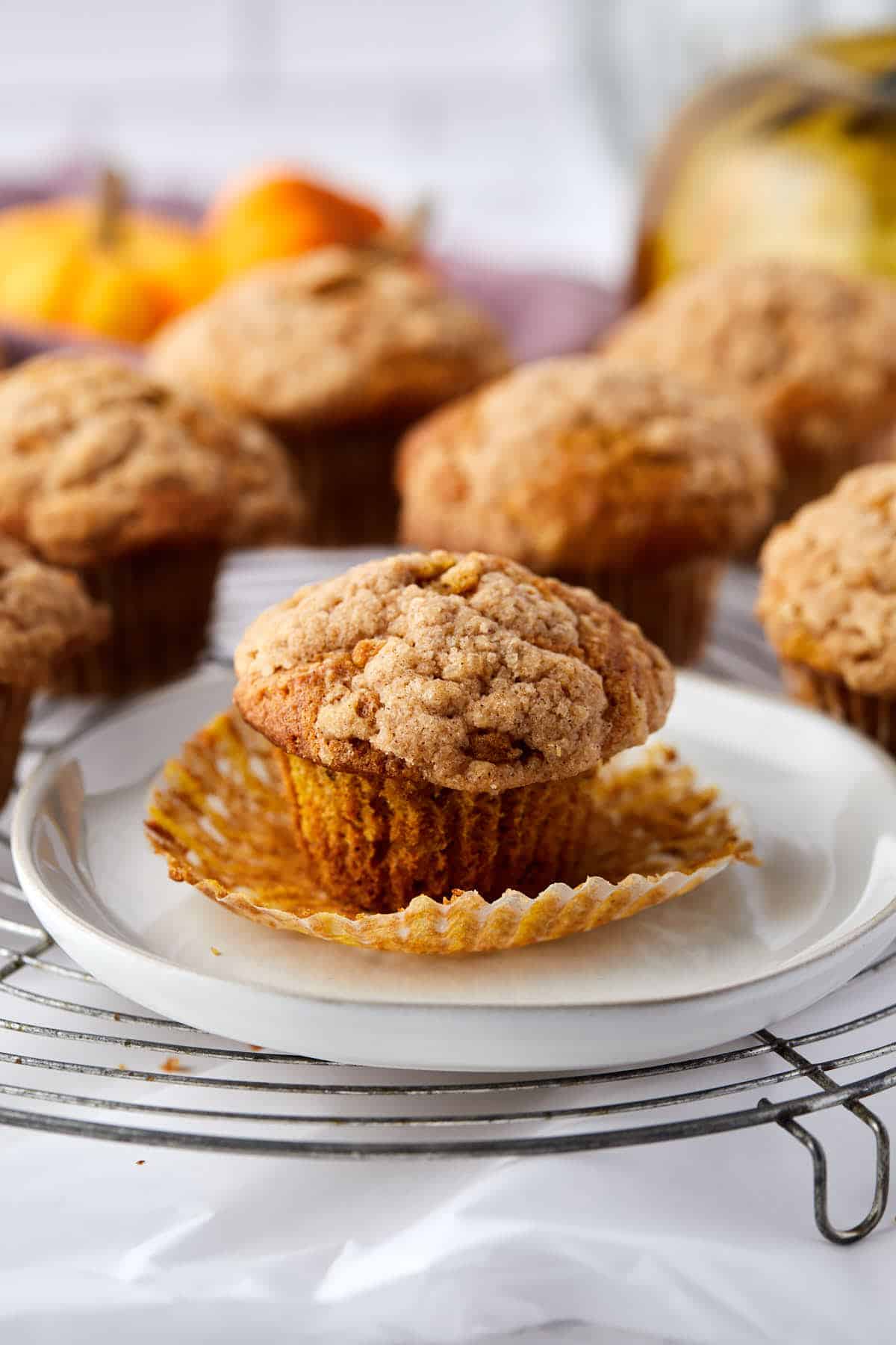 A freshly baked muffin with a crumbly top sits on a plate with its wrapper peeled open. More muffins are on a cooling rack in the background, with pumpkins faintly visible, suggesting an autumn theme.