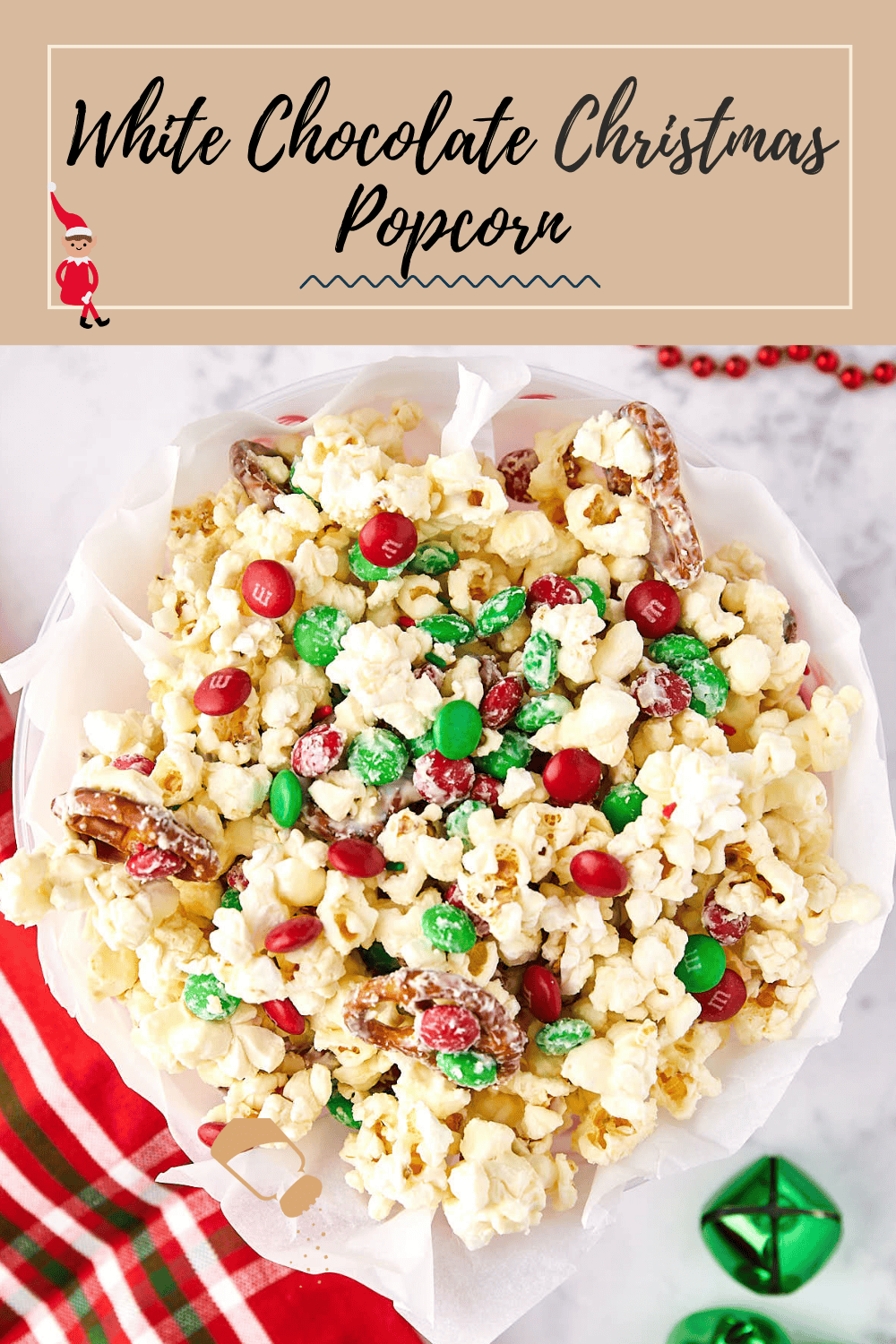 A festive bowl of white chocolate popcorn is mixed with red and green candies and pretzels. A red and white striped cloth and decorative bells are next to the bowl, creating a holiday-themed setting.