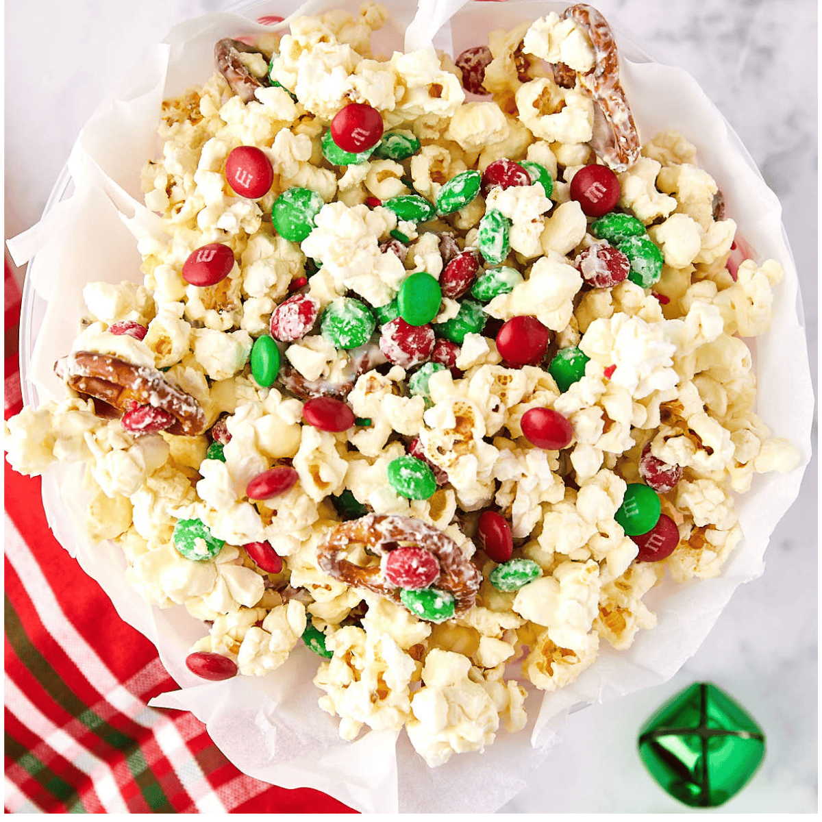 A festive bowl of popcorn mixed with red and green candies and pretzels, topped with white chocolate drizzle. It is placed on a white surface, and a red and green striped cloth and a green jingle bell are nearby.