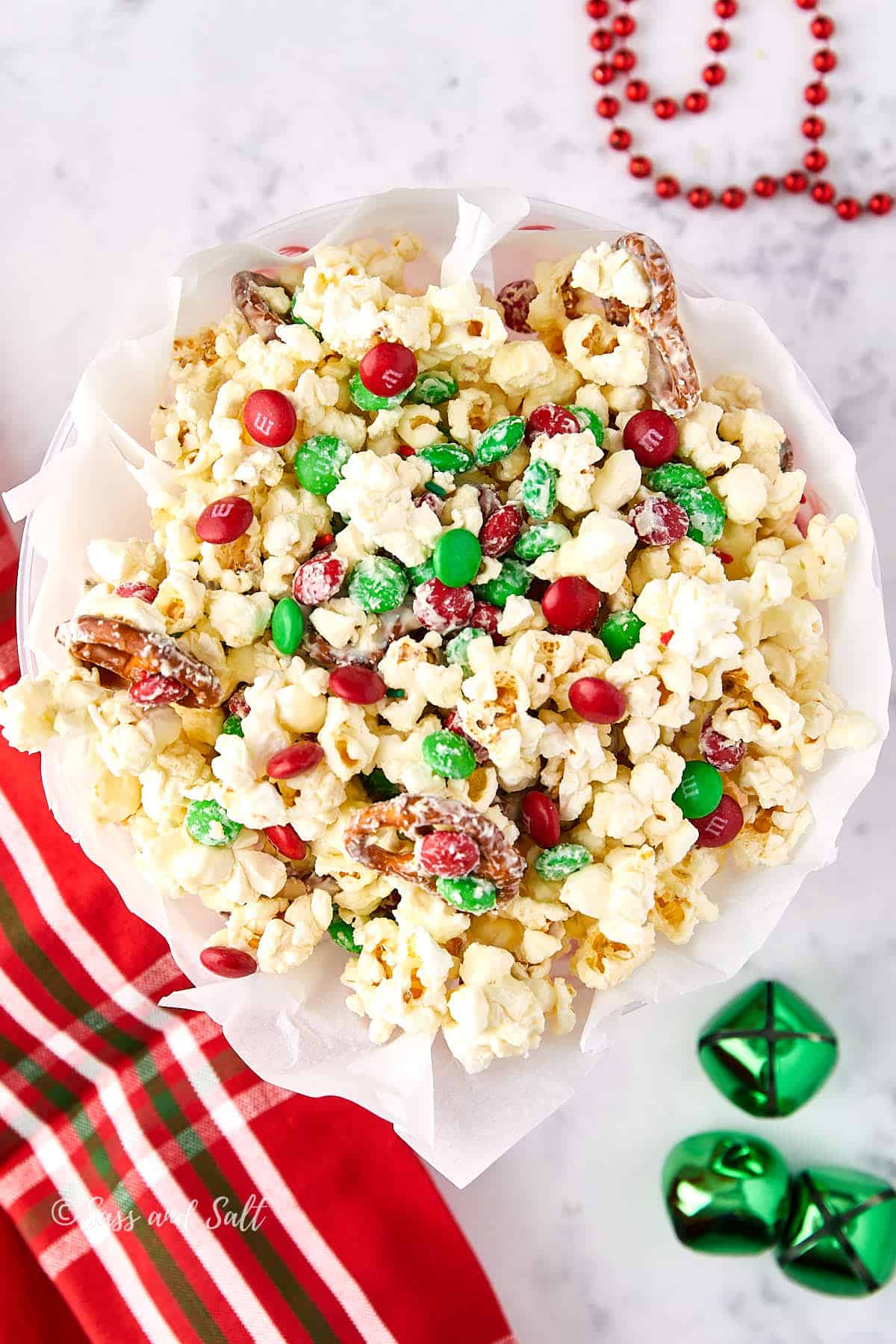 A festive bowl of popcorn mixed with red and green candies and pecans, placed on a white surface. Red and green jingle bells and a striped red cloth are nearby, adding to the holiday theme.