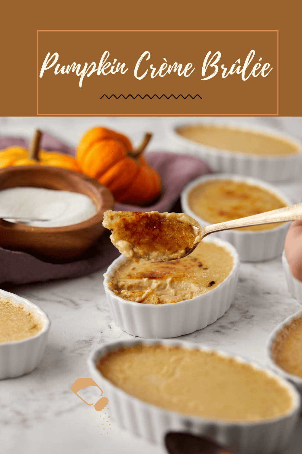 A spoon scoops creamy pumpkin crème brûlée from a ramekin. Several ramekins filled with the dessert are visible, set on a marble surface. Mini pumpkins and a wooden bowl of sugar are in the background. A "Pumpkin Crème Brûlée" label is at the top.