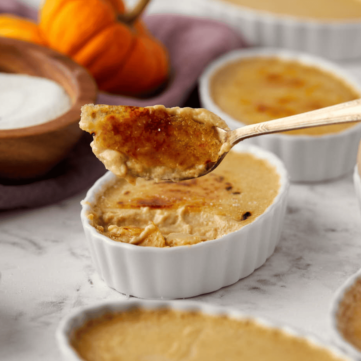 A spoon filled with creamy caramelized pumpkin crème brûlée hovers over a white ramekin on a marble surface. In the background, a small pumpkin and a wooden bowl of sugar can be seen.