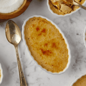 Oval-shaped crème brûlée in a white ramekin, topped with a caramelized sugar crust. A vintage spoon lies nearby. Other ramekins and a wooden bowl of sugar are partially visible on a marble surface.