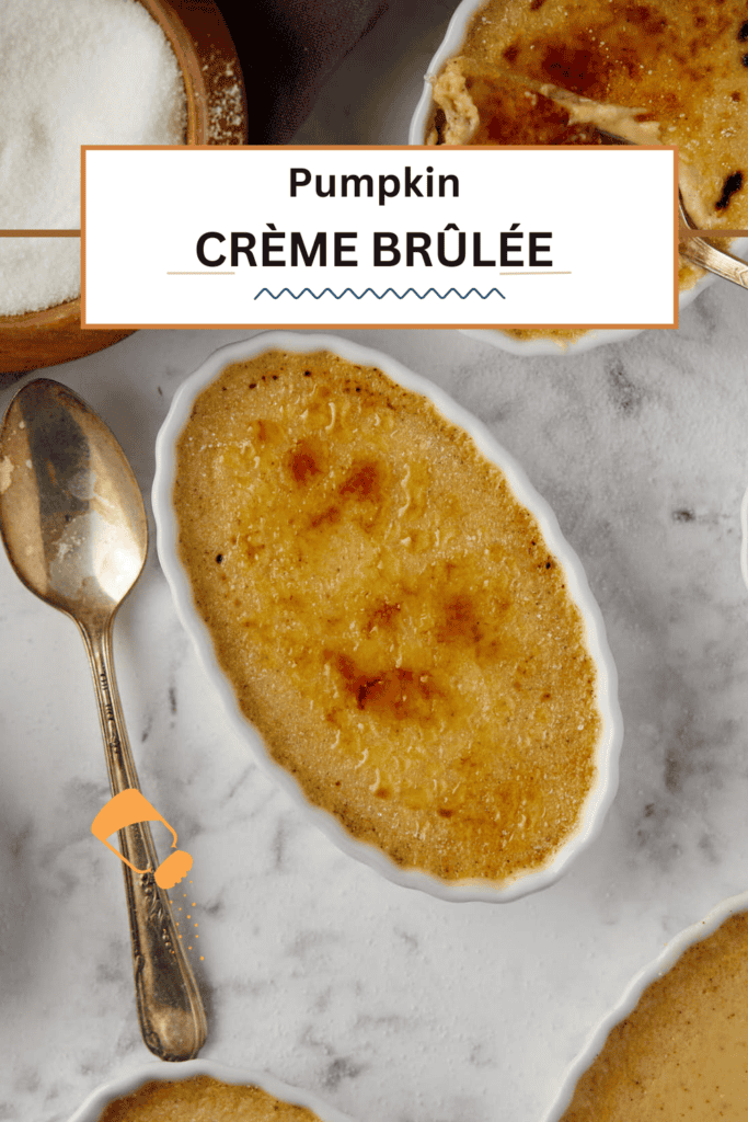 Top view of a pumpkin crème brûlée in an oval dish with a golden-brown, caramelized surface. It is placed on a marble countertop, accompanied by a vintage spoon and a bowl of sugar in the corner. A label reads "Pumpkin Crème Brûlée.