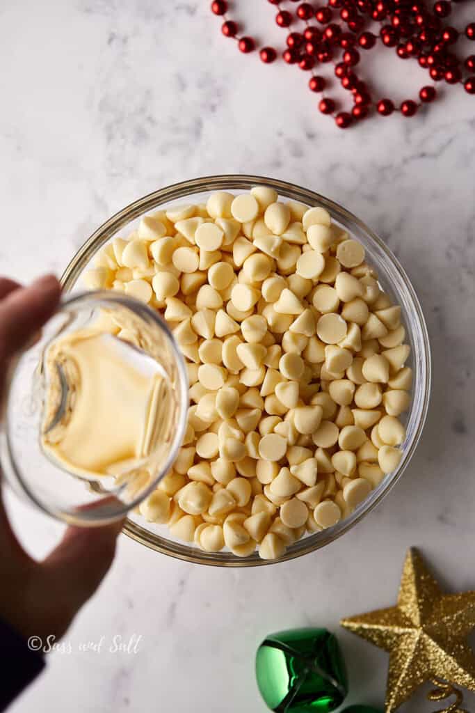 A glass bowl filled with white chocolate chips on a marble surface, with a hand pouring coconut oil from a small bowl. Red beads and a decorative star and bell are partially visible.