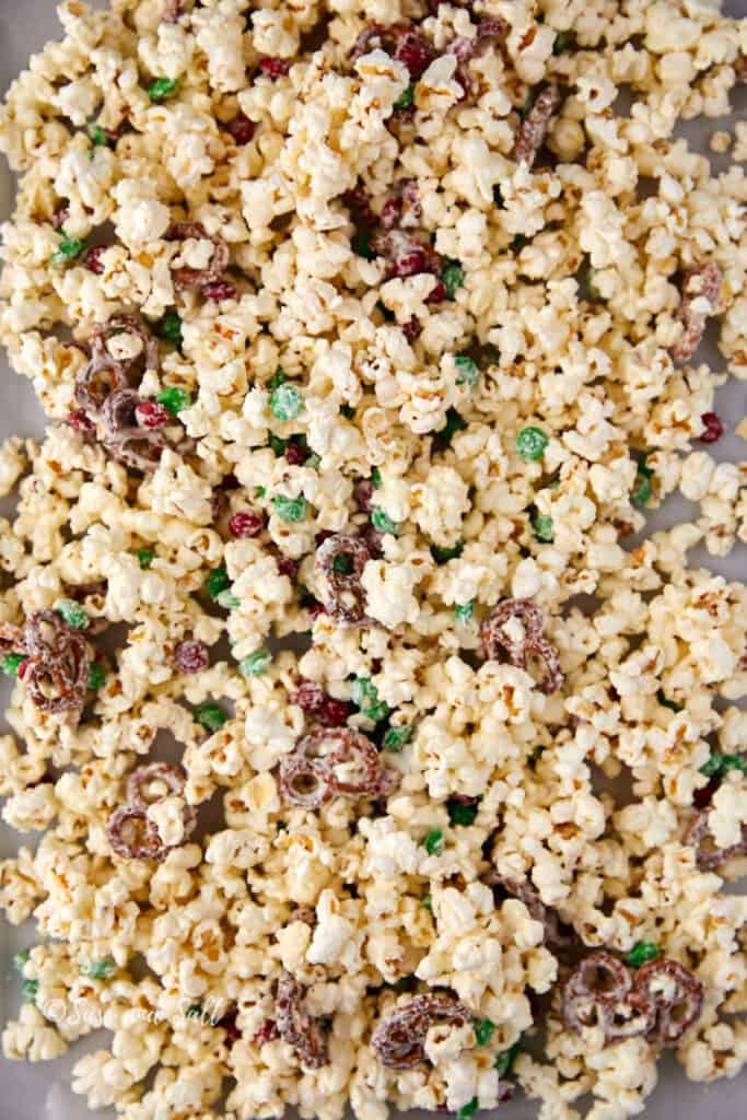 A close-up of popcorn mixed with chocolate-coated pretzels and colorful candy-coated chocolate pieces, spread on a flat surface.