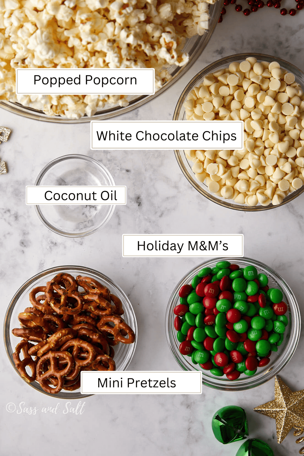 A marble surface holds bowls filled with popped popcorn, white chocolate chips, coconut oil, holiday M&M's, and mini pretzels. Gold stars and red beads are scattered around for decoration.