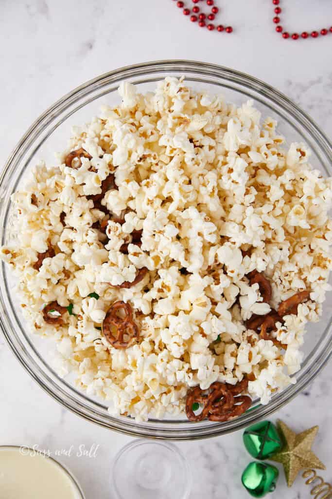 A bowl of popcorn mixed with pretzels and green candies is on a marble surface. Nearby are festive decorations, including a gold star and red beads.