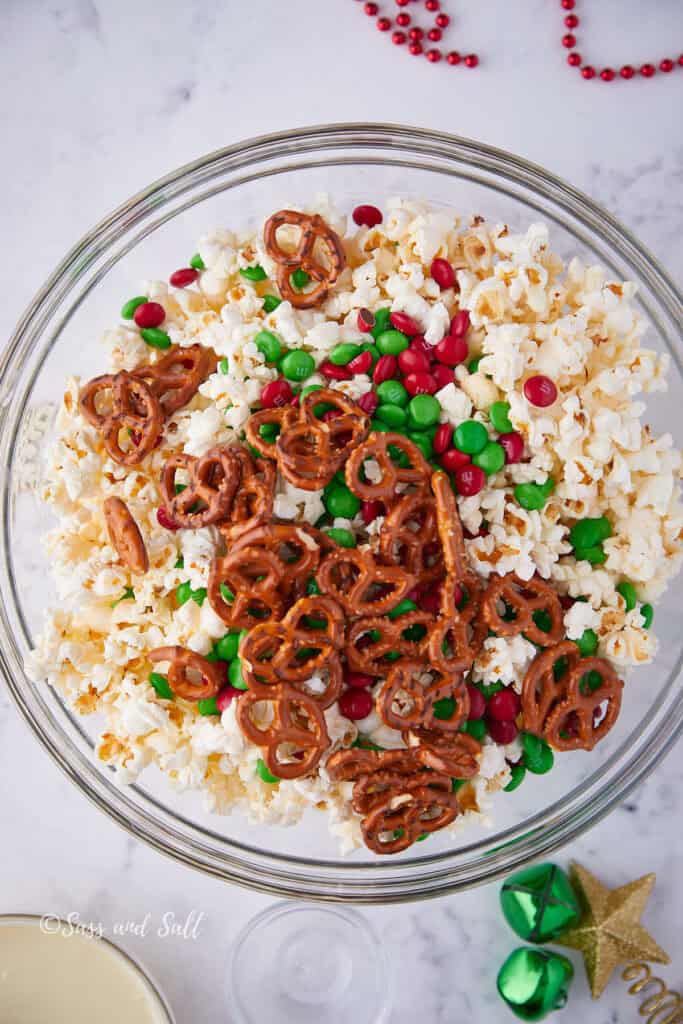 A glass bowl filled with popped popcorn, red and green candy-coated chocolates, and pretzels on a white marble surface. Decorative Christmas ornaments are nearby.