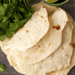 A stack of flour tortillas is placed on a wooden surface, accompanied by fresh cilantro and a small bowl containing lime wedges.