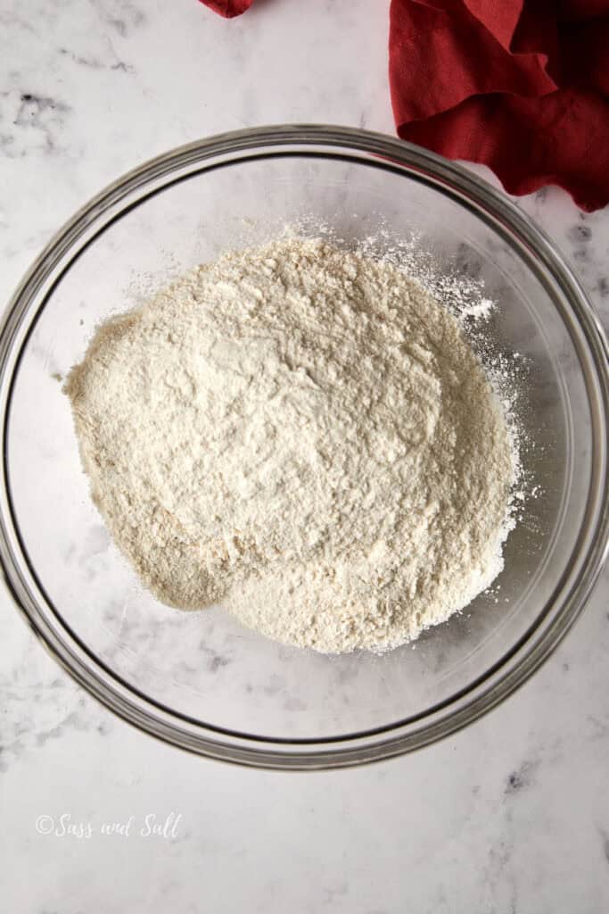 A glass bowl filled with flour sits on a marble countertop. There's a red cloth partially visible in the top right corner.