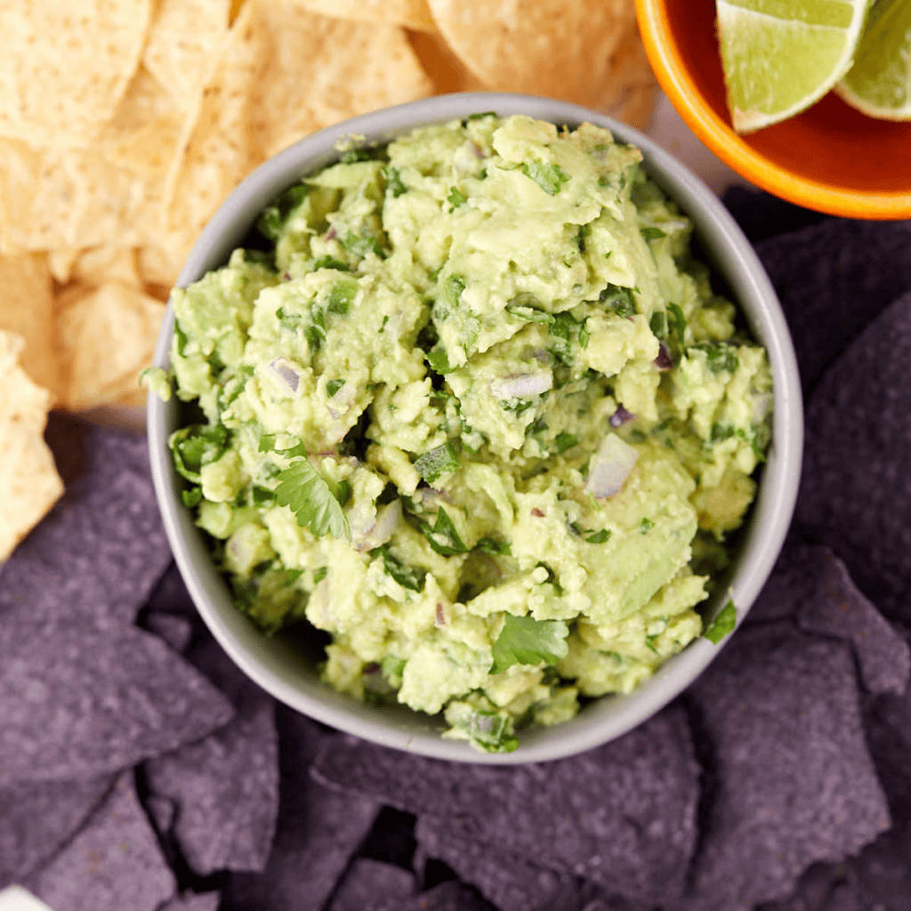 A bowl of guacamole garnished with cilantro and diced onions is surrounded by blue and yellow tortilla chips. Lime wedges are visible in a small bowl beside the guacamole.