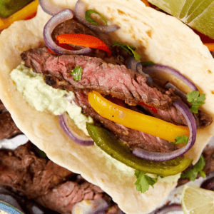 Close-up of a delicious steak fajita with. steak slices, colorful bell peppers, red onion rings, and a dollop of guacamole on a warm tortilla. Sprigs of cilantro garnish the taco, and lime wedges are visible nearby.