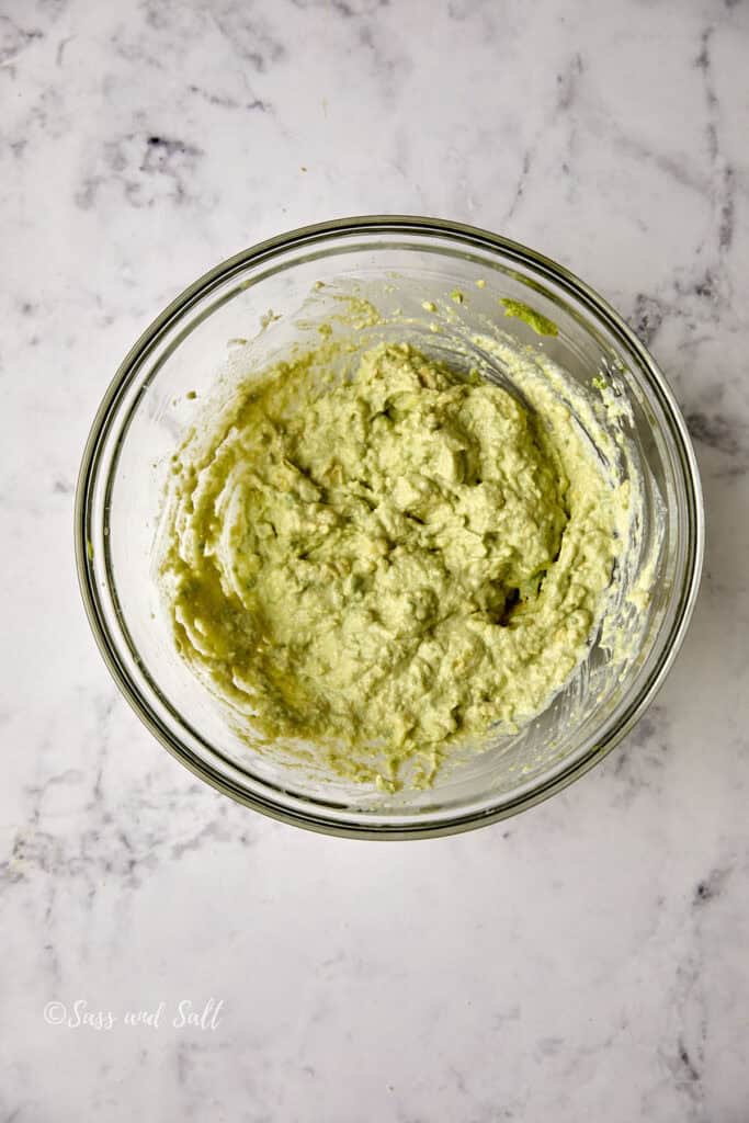 A clear glass bowl filled with mashed avocado sits on a white marble countertop. The creamy mixture has a smooth texture and subtle green color.