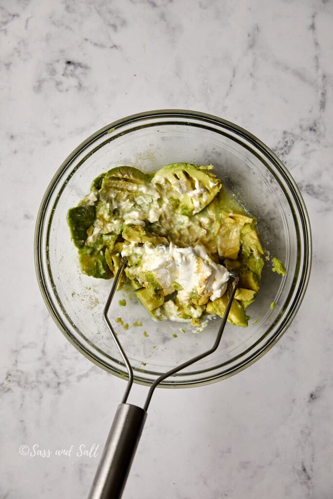 A glass bowl on a marble surface contains mashed avocado mixed with sour cream, with a metal masher resting inside.