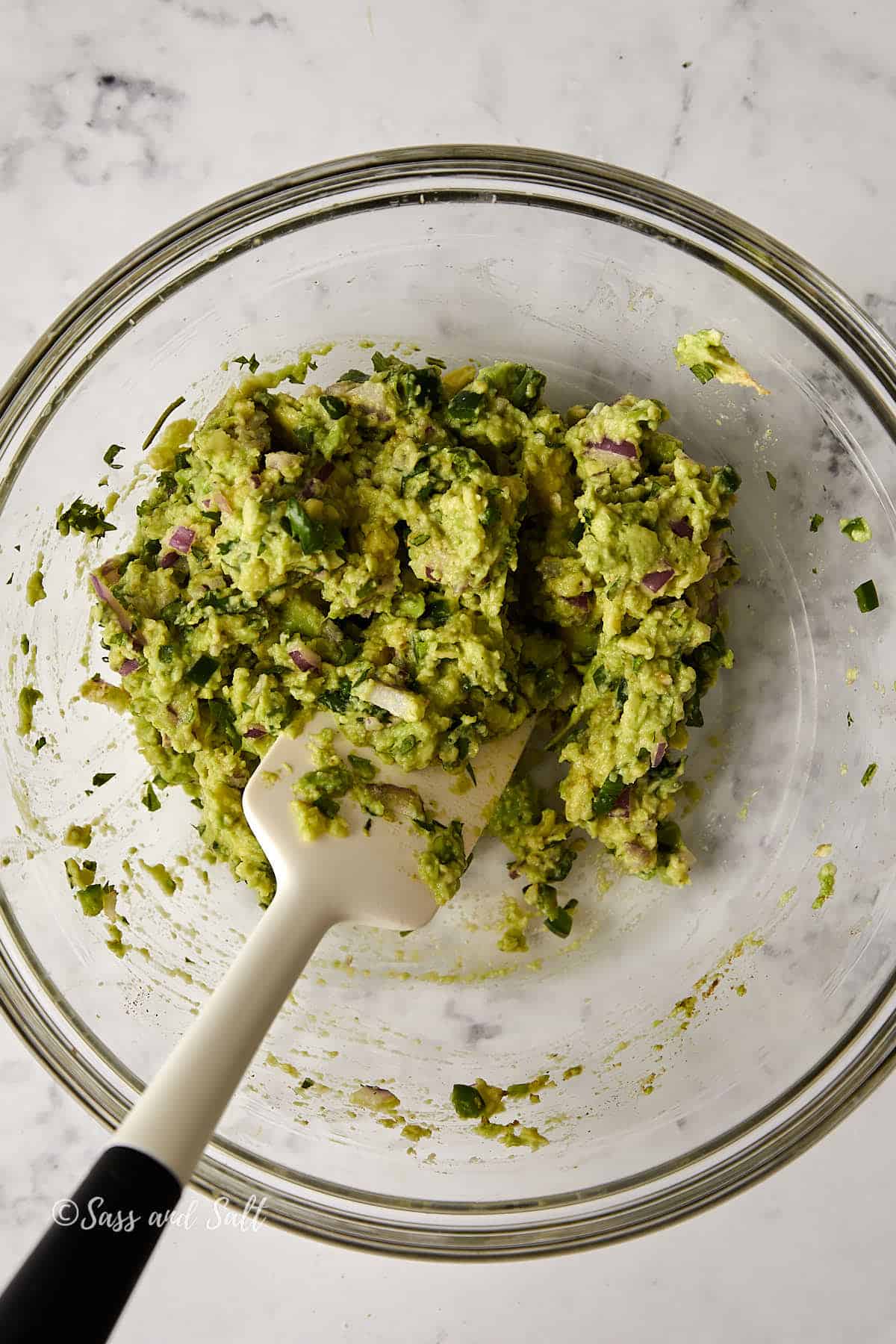 A clear glass bowl filled with chunky guacamole, featuring visible pieces of avocado, chopped red onion, and cilantro. A white spatula rests in the bowl on a marble countertop.