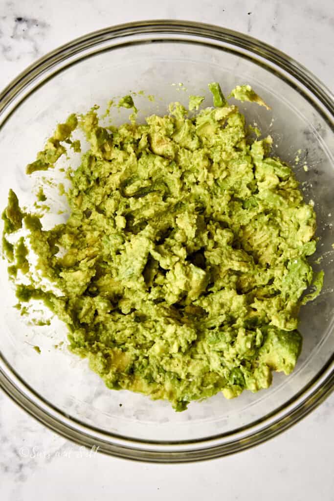 A clear glass bowl filled with creamy, mashed avocado sits on a marble countertop. The avocado is bright green and smoothly blended, indicating preparation for a dish like guacamole.