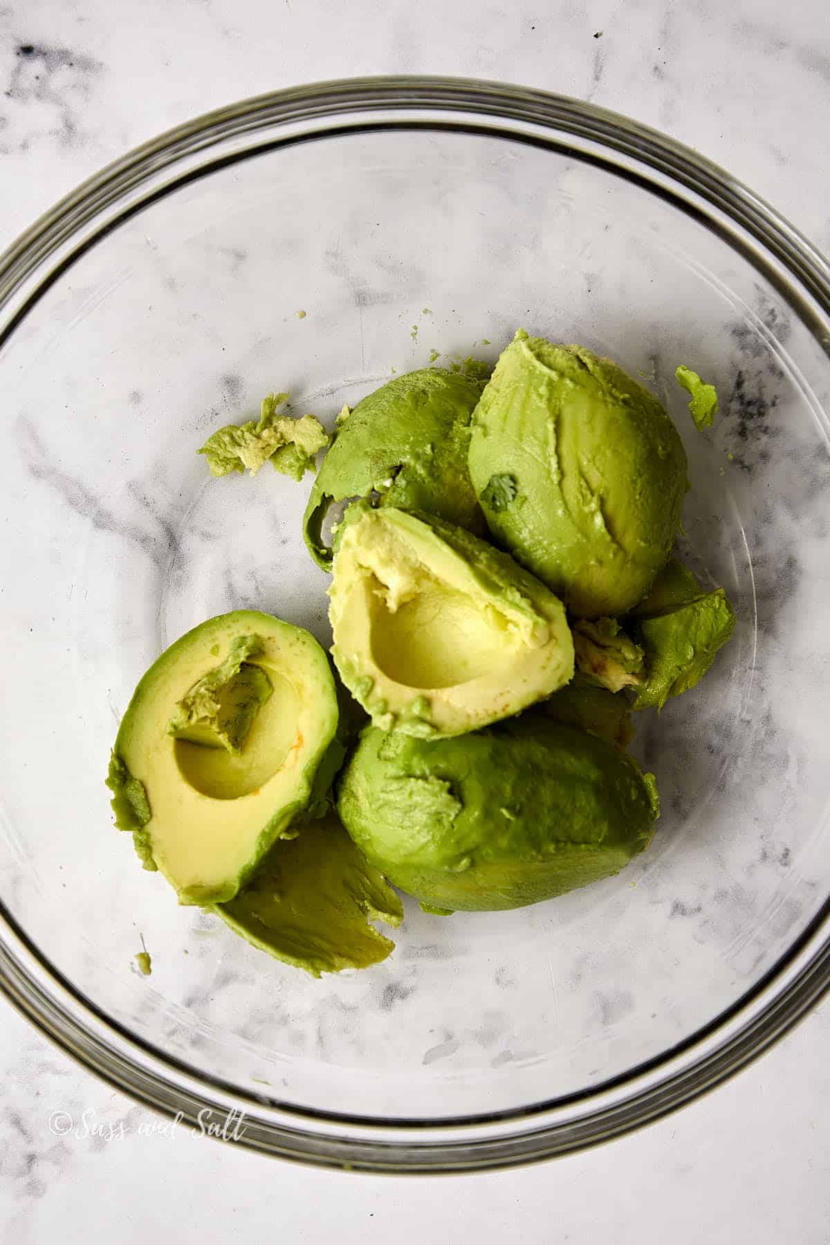 Halved avocados with pits removed in a glass bowl set on a marble countertop.