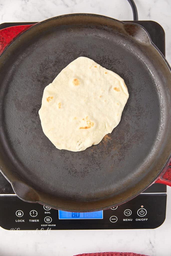 A flatbread is cooking on a cast iron skillet placed on an electric stovetop with a digital display. The stovetop is black with red handles, and the surrounding surface is white marble.