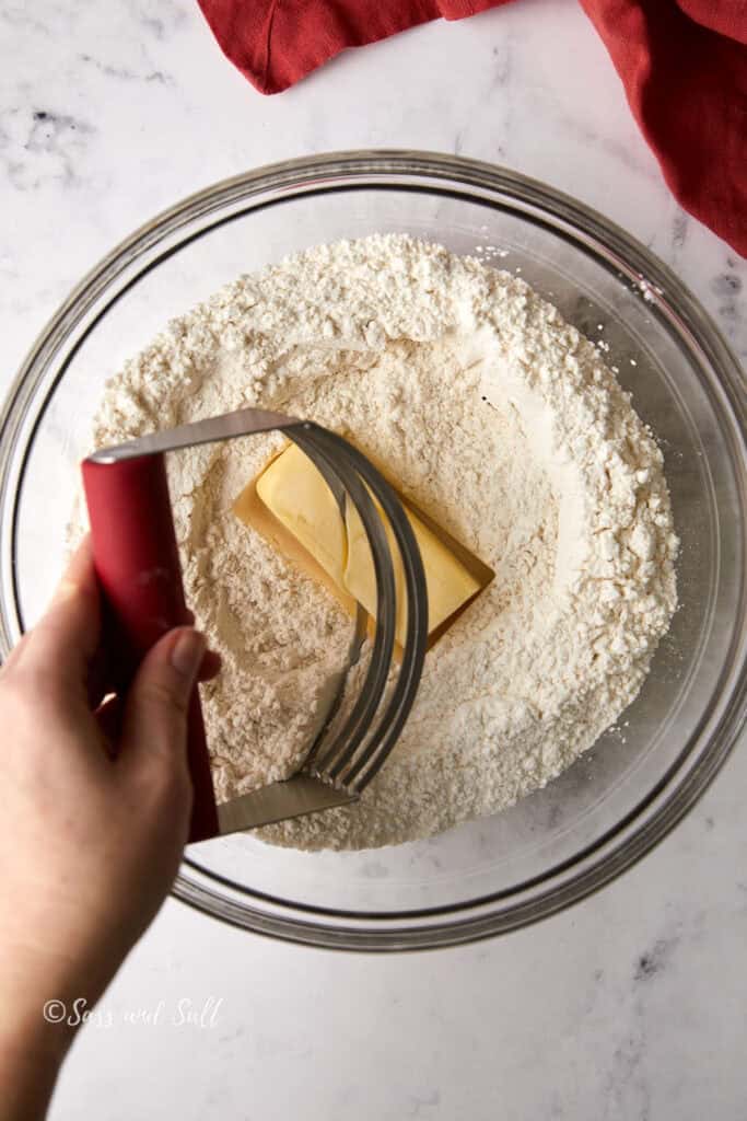 A person uses a pastry cutter to mix a block of butter into a bowl of flour. The bowl is transparent and rests on a white marble surface. A red cloth is partially visible in the top corner.