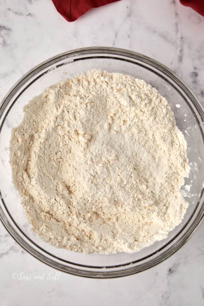 A glass bowl filled with flour mixed with butter marble countertop. A red cloth is partially visible in the top left corner.