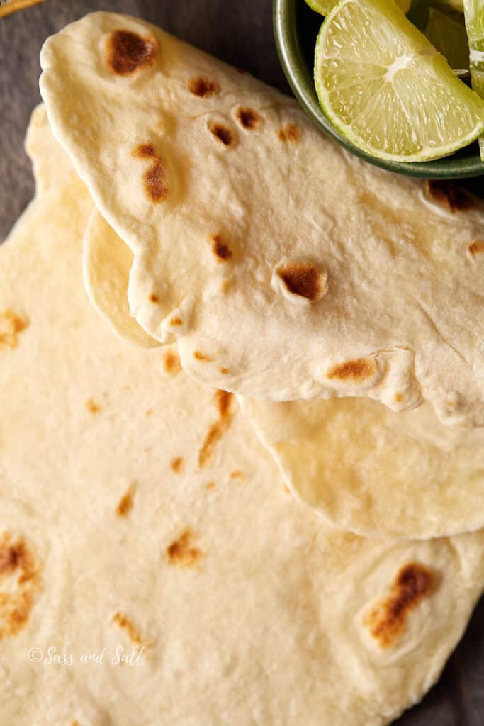 Flatbreads stacked on a wooden surface next to a small bowl filled with lime wedges.