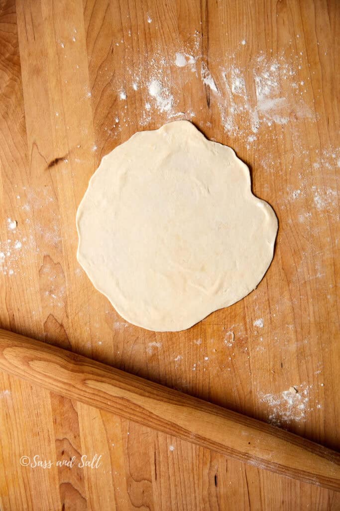 A round piece of rolled-out dough on a floured wooden surface, with a wooden rolling pin nearby.