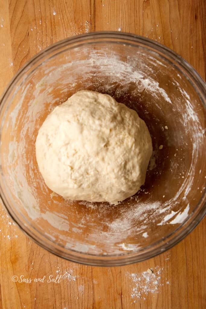 A ball of dough sits in a clear glass bowl on a wooden surface, lightly dusted with flour.