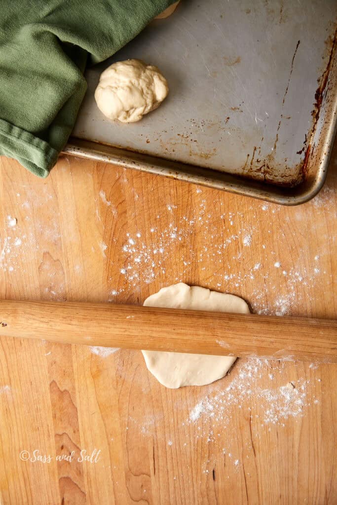 A wooden rolling pin rolls out dough on a floured wooden surface. A green cloth lies on a metal baking sheet in the background, next to a small ball of dough.