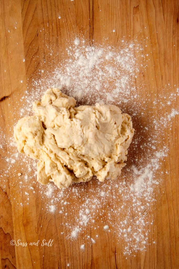 Raw dough on a wooden surface with scattered flour around it. The texture appears rough, and the wood has a natural grain pattern.