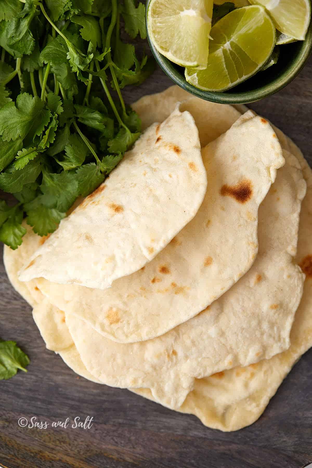 A top view of freshly made tortillas stacked on a wooden surface, accompanied by a bowl of lime wedges and a bunch of fresh cilantro.