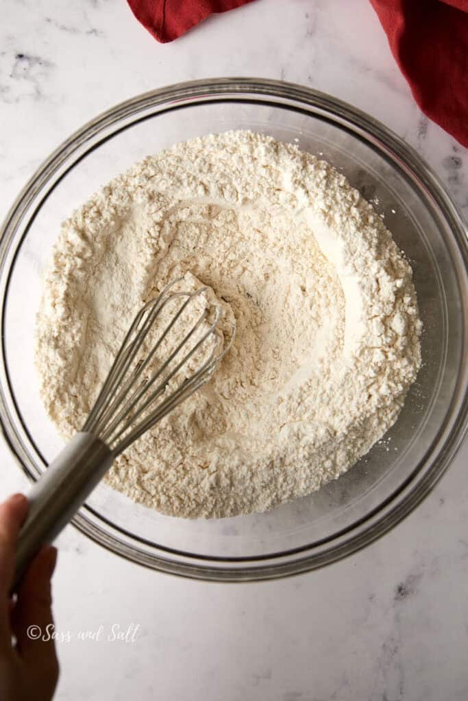 A hand using a whisk to mix flour in a clear glass bowl, set on a marble countertop. A red cloth is partially visible in the corner.