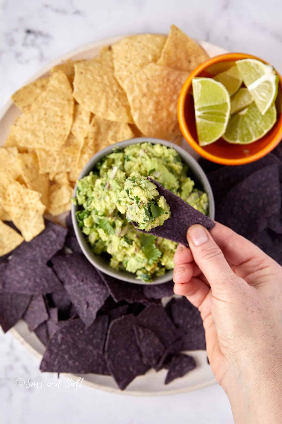 A hand holds a blue corn chip with guacamole. The plate features more blue and yellow corn chips, with a bowl of guacamole in the center. A smaller bowl with lime wedges is placed nearby.