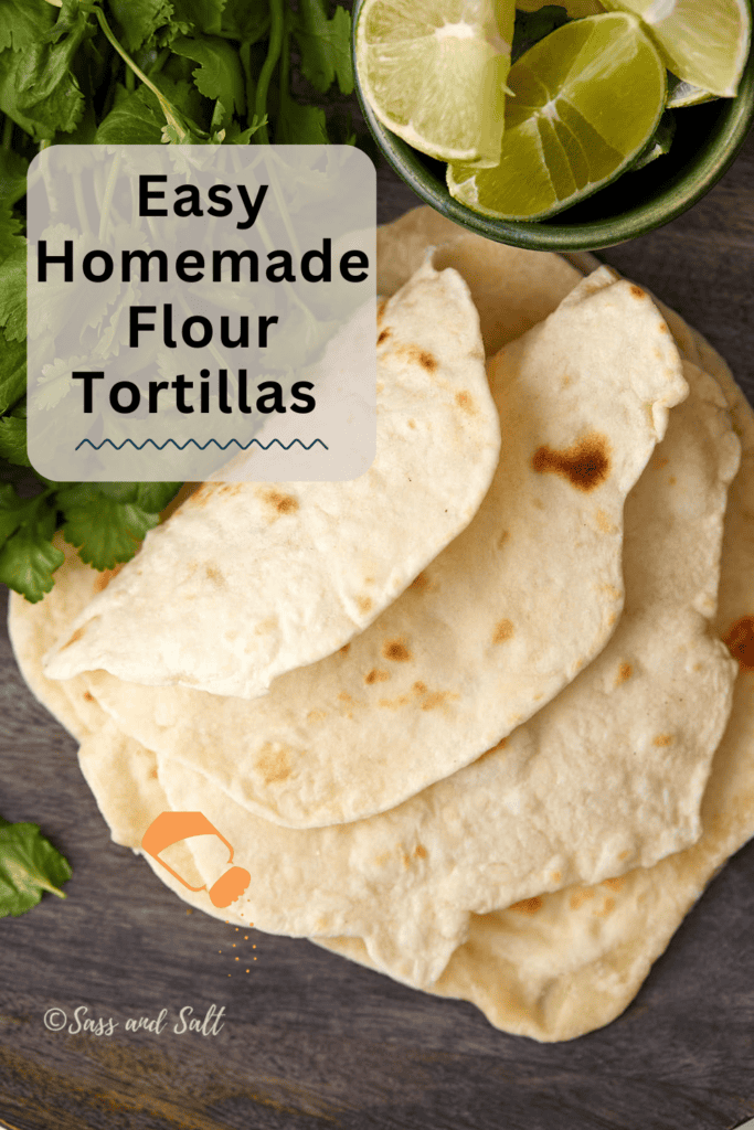 Top view of homemade flour tortillas stacked on a wooden surface, garnished with cilantro and lime wedges in a small green bowl. Text overlay reads "Easy Homemade Flour Tortillas.