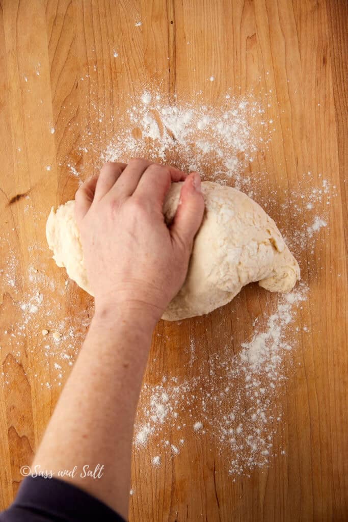 A person's hand kneading dough on a floured wooden surface. The dough is light in color and slightly dusted with flour, surrounded by scattered flour on the table.