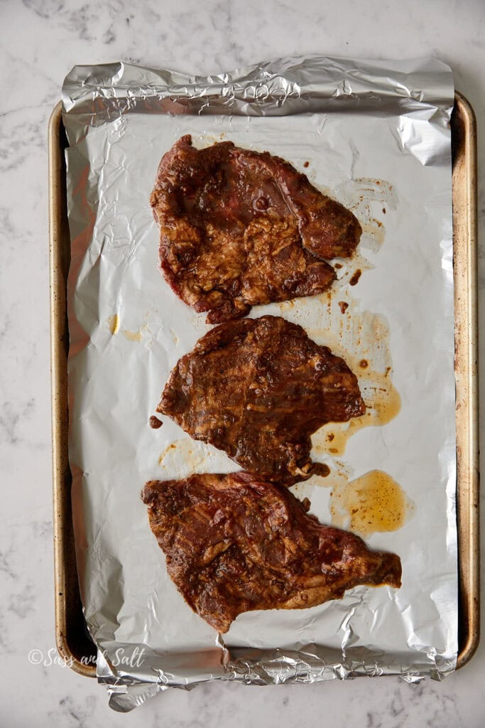 Three marinated steaks on a foil-lined baking sheet, ready for cooking. The steaks are seasoned and arranged in a row. The background is a marble countertop.