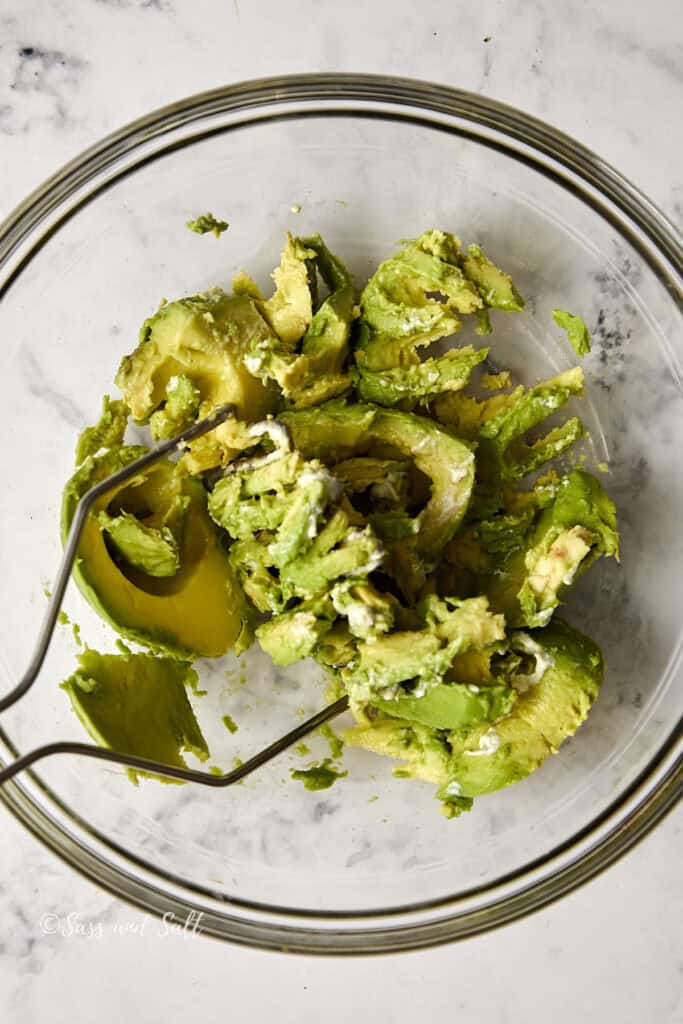 A glass bowl containing roughly mashed avocados. A metal masher rests inside the bowl, surrounded by chunks of avocado. The setting is on a light marble surface.