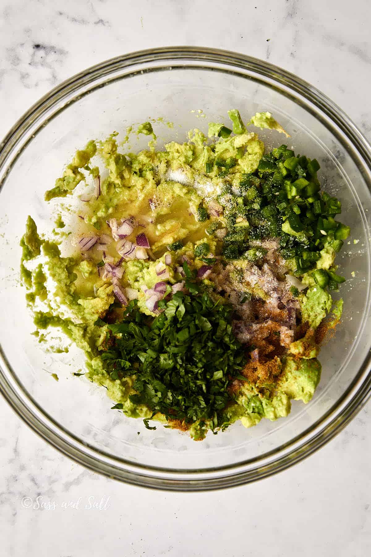 A glass bowl on a marble surface contains a mixture of chopped green herbs, diced red onions, jalapeños, and mashed avocado, likely ingredients for guacamole. Seasonings are also visible sprinkled on top.