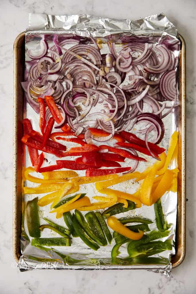 A baking tray lined with foil holds sliced red onions, red bell peppers, yellow bell peppers, and green bell peppers. The vegetables are arranged in rows, ready for roasting.