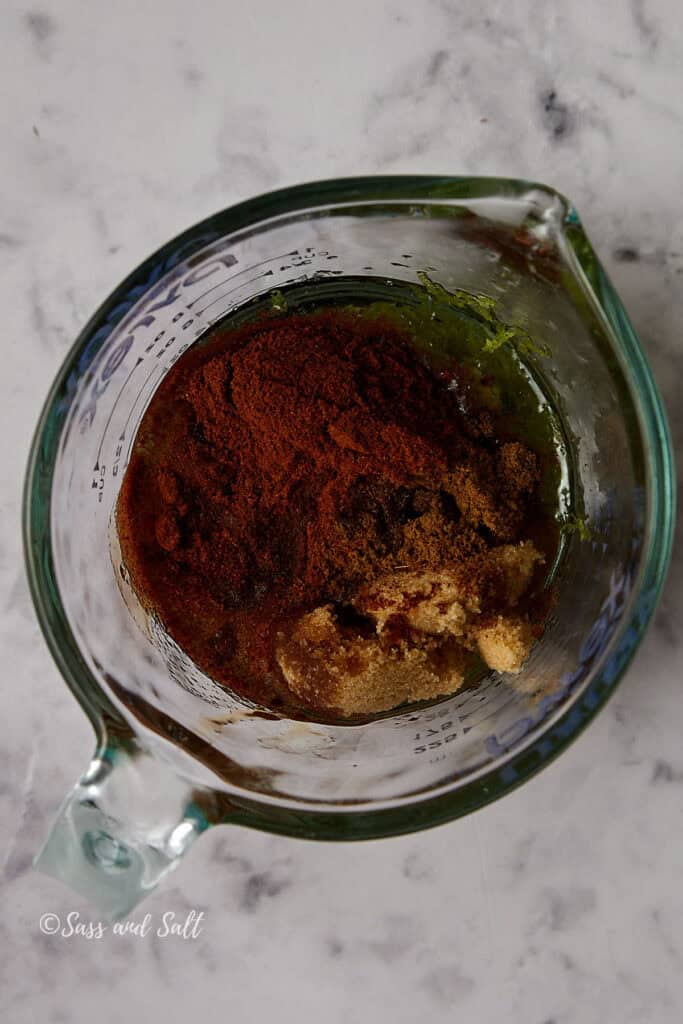 A glass measuring cup on a marble surface contains a mixture of spices, brown sugar, and dark liquid. The blend appears ready for mixing with other ingredients.