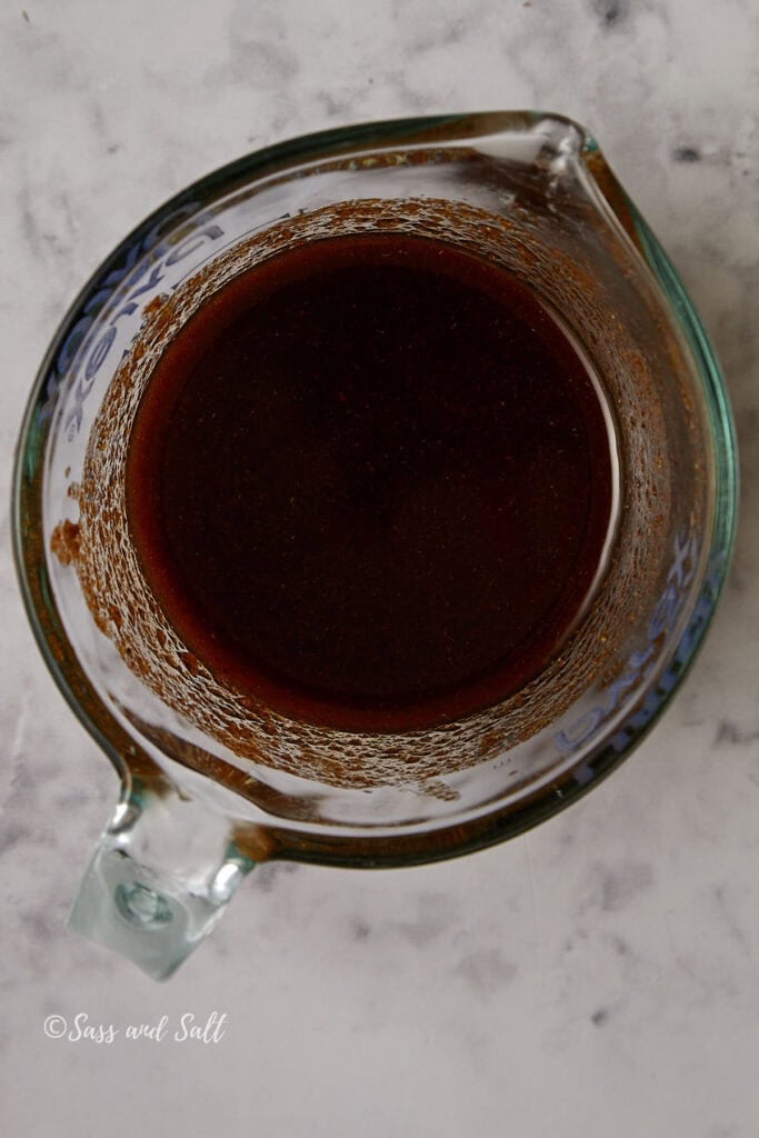 A glass measuring cup filled with dark brown liquid sits on a marbled countertop. The contents appear smooth, with traces of residue around the sides of the cup. On the bottom left corner, faint text reads "Sass and Salt.