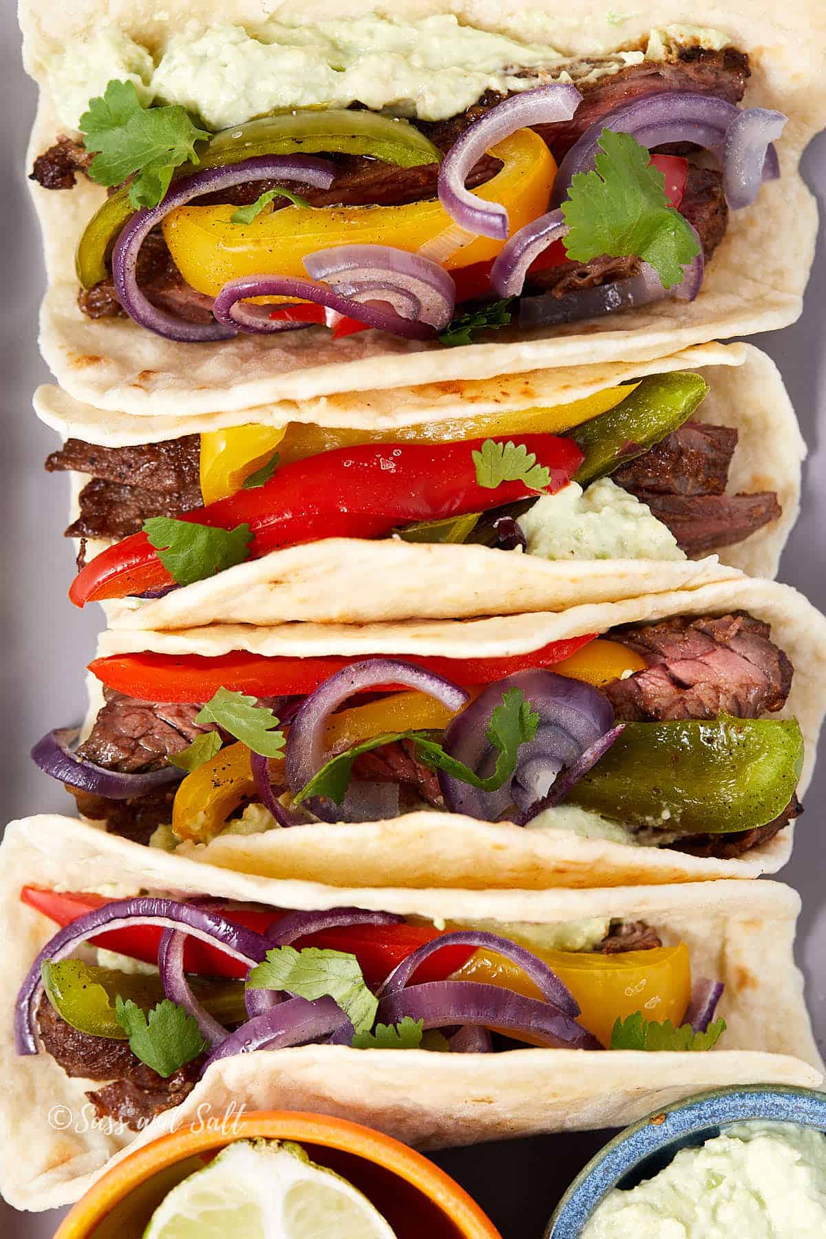 Three soft tacos filled with slices of steak, colorful bell peppers, red onions, and cilantro, topped with guacamole. A small bowl of guacamole and a lime wedge are visible next to the tacos.
