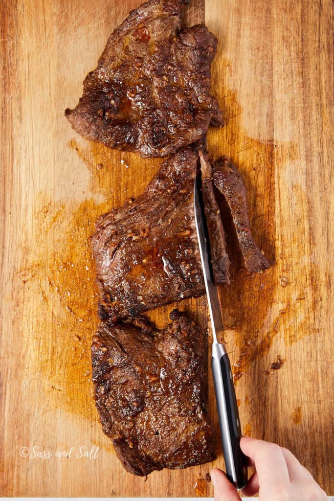 A hand slicing cooked steak on a wooden cutting board. The steak is glistening with juices, and the wood has a slightly worn texture. The meat is partially cut into thick pieces, and a knife is being used to make the slices.