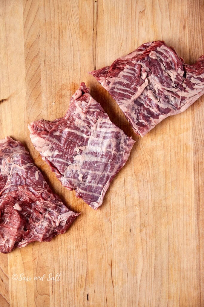 Three pieces of raw beef short ribs are placed on a wooden cutting board. The meat is marbled and unseasoned, showing distinct texture and grain.