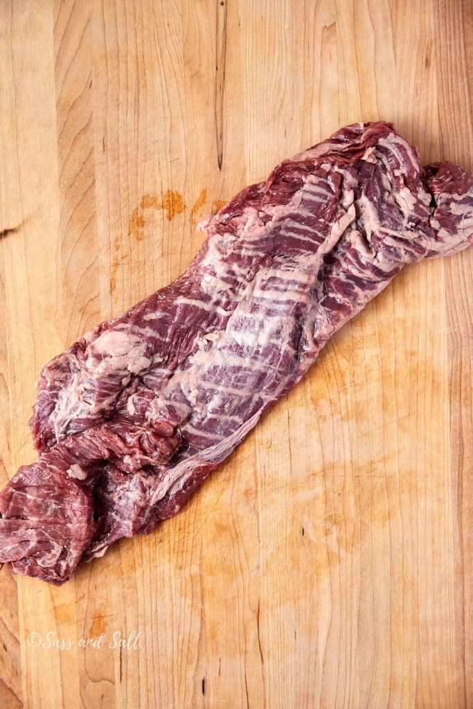 A raw piece of beef skirt steak is laid out on a wooden cutting board. The marbling and texture of the meat are visible, with some fat lining the edges. The board has a natural wood grain pattern.