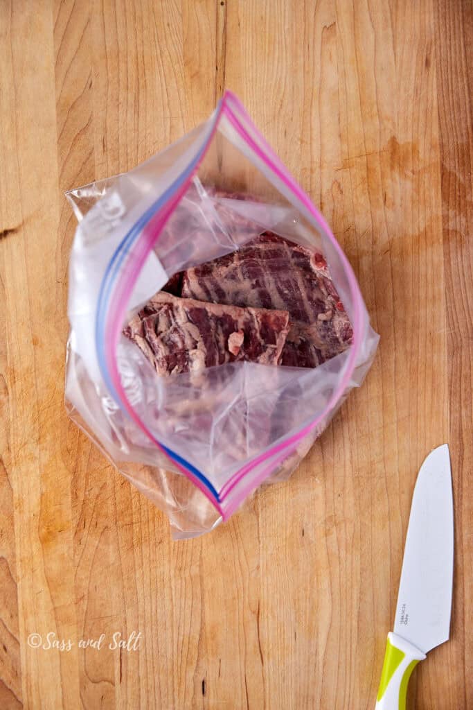 A clear plastic bag containing meat lies on a wooden surface next to a white-handled kitchen knife.